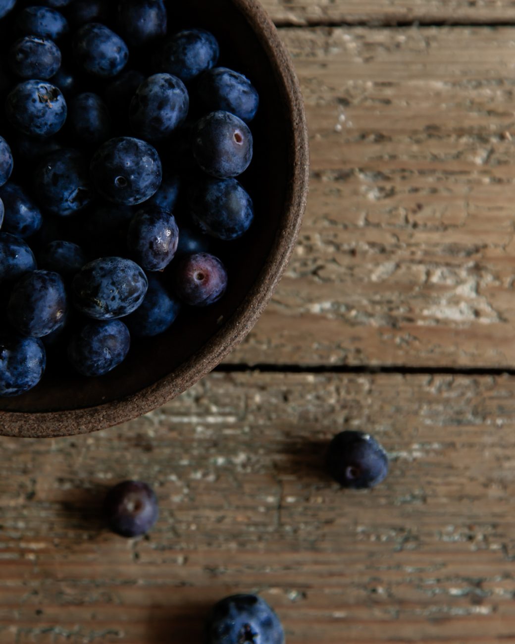 A wooden bowl filled with blueberries sits on a rustic wooden surface. A few blueberries have spilled out of the bowl and lie on the table next to it. The blueberries are dark and plump, contrasting with the textured, weathered wood of the table. The image has a cozy, natural feel with a focus on the fresh berries.