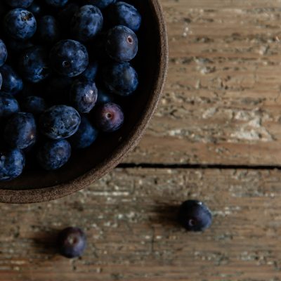 A wooden bowl filled with blueberries sits on a rustic wooden surface. A few blueberries have spilled out of the bowl and lie on the table next to it. The blueberries are dark and plump, contrasting with the textured, weathered wood of the table. The image has a cozy, natural feel with a focus on the fresh berries.