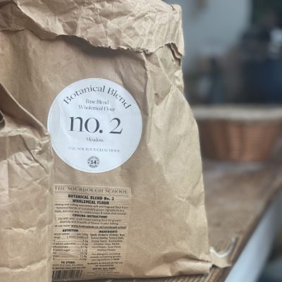 The image shows a large brown paper bag labeled "Botanical Blend no. 2" from The Sourdough School. The bag appears slightly crumpled, giving it a natural, handcrafted look. The label includes text indicating that it is a blend of wholegrain flours. The bag sits on a wooden countertop, and the background is softly blurred.