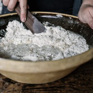 A close-up of hands mixing ingredients in a large, white mixing bowl. The person is using a wooden spoon to stir the mixture, which appears to be a batter or dough. The bowl is on a wooden surface, and the background is blurred, keeping the focus on the hands and the mixing process.