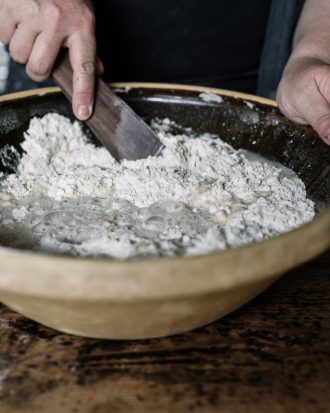A close-up of hands mixing ingredients in a large, white mixing bowl. The person is using a wooden spoon to stir the mixture, which appears to be a batter or dough. The bowl is on a wooden surface, and the background is blurred, keeping the focus on the hands and the mixing process.