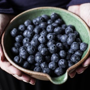 A person is holding a bowl filled with fresh blueberries. The bowl is green on the inside and has a rustic, earthy brown exterior. The blueberries are plump and have a deep blue hue, with a slightly frosted appearance. The person's hands gently cradle the bowl, and the background is blurred, focusing attention on the berries and the bowl.