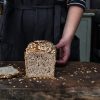 The image shows a loaf of artisanal sourdough bread sliced open to reveal a beautifully airy and open crumb structure. The crust is golden brown a with oat flakes scattered on the top. The inside of the bread appears soft and slightly glossy, with a network of irregular holes typical of well-fermented sourdough. The background is a simple, neutral surface, allowing the focus to remain on the bread’s texture and inviting appearance.