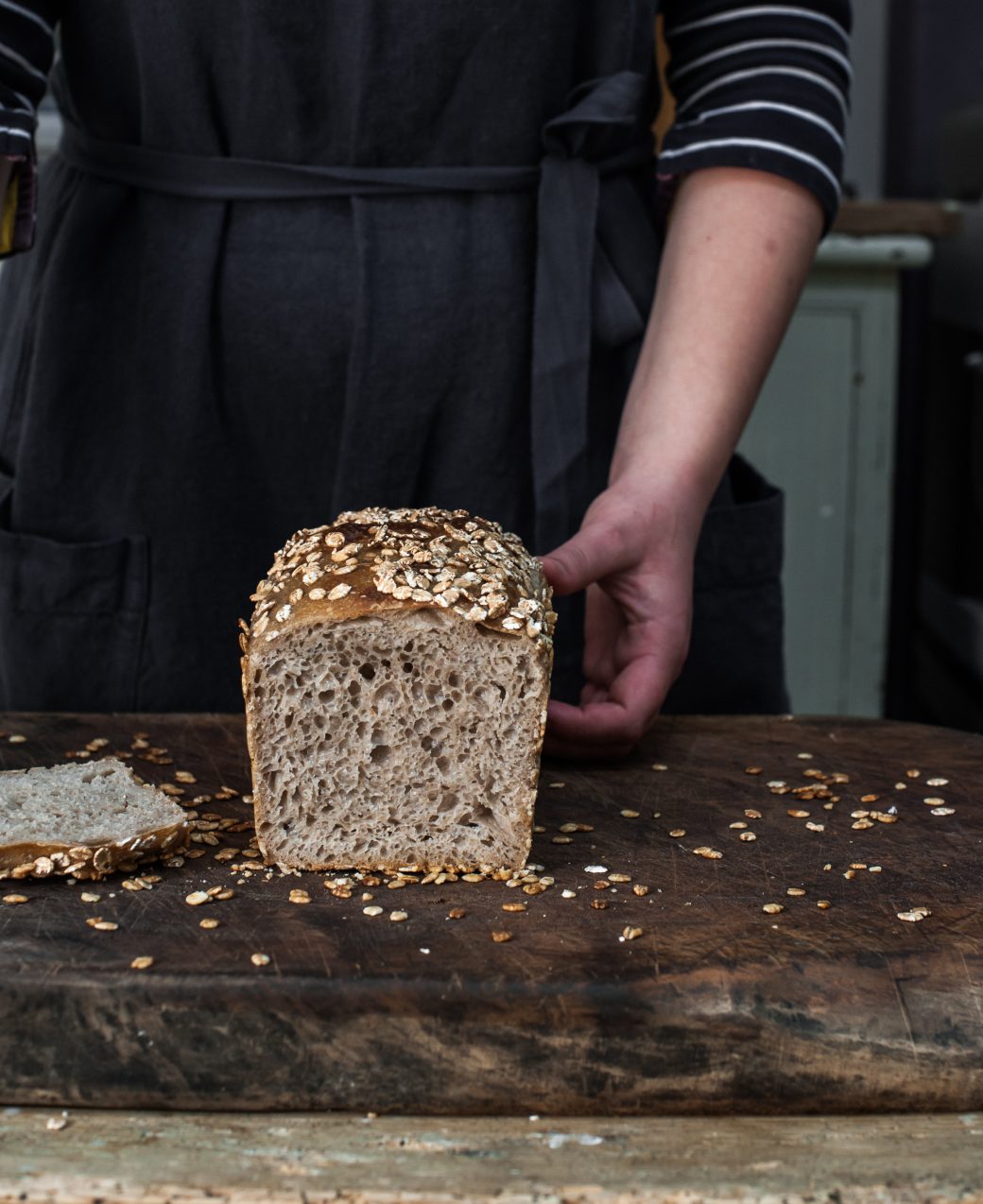 The image shows a loaf of artisanal sourdough bread sliced open to reveal a beautifully airy and open crumb structure. The crust is golden brown a with oat flakes scattered on the top. The inside of the bread appears soft and slightly glossy, with a network of irregular holes typical of well-fermented sourdough. The background is a simple, neutral surface, allowing the focus to remain on the bread’s texture and inviting appearance.