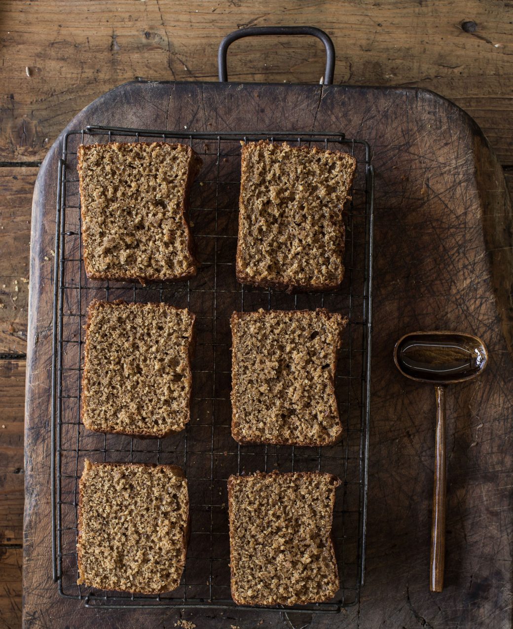 Six slices of cake on a cooling rack on top of a wooden board, with a spoon filled with honey beside.