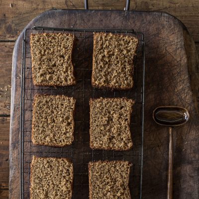 Six slices of cake on a cooling rack on top of a wooden board, with a spoon filled with honey beside.