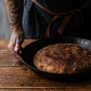 A tattooed arm holding a handle to a baking dish with a golden miso flatbread inside