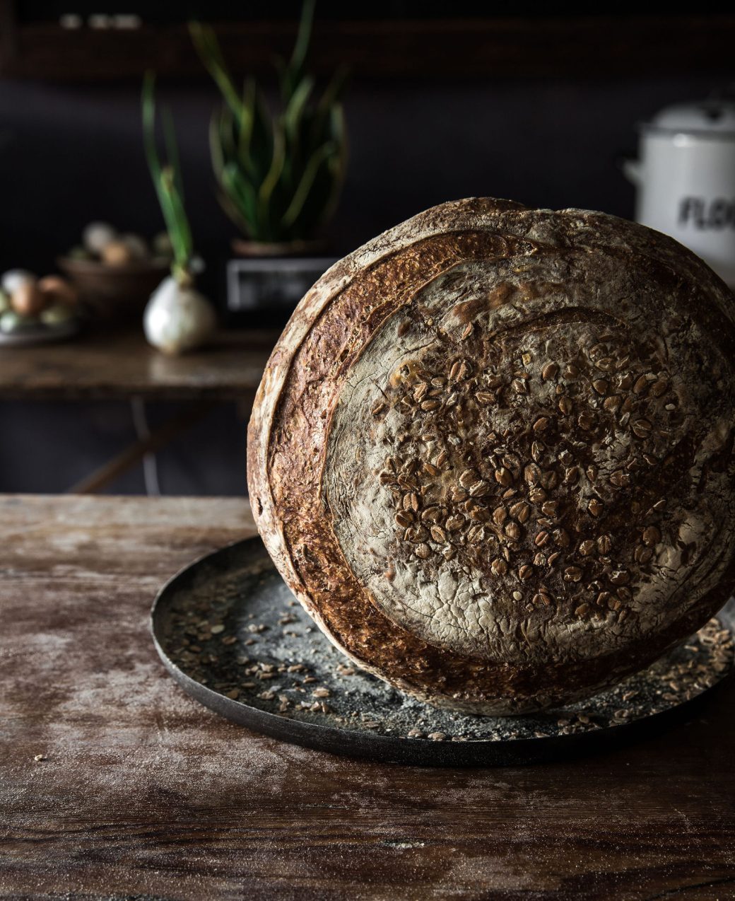 A beer boule topped with barley flakes is standing on its side on top of a black plate placed on a wooden table.